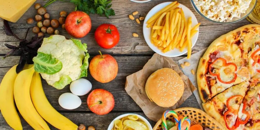Mesa con alimentos variados: plátanos, huevos, verduras, pizza, hamburguesa y patatas fritas