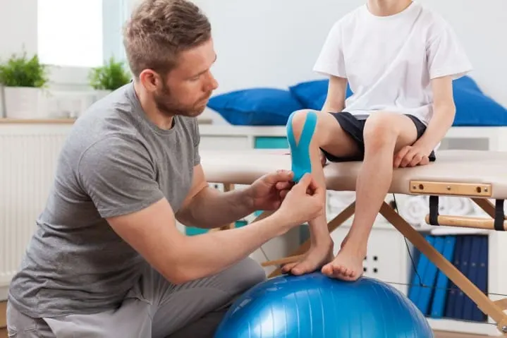Fisioterapeuta colocando un vendaje en la rodilla de un niño sentado sobre una pelota