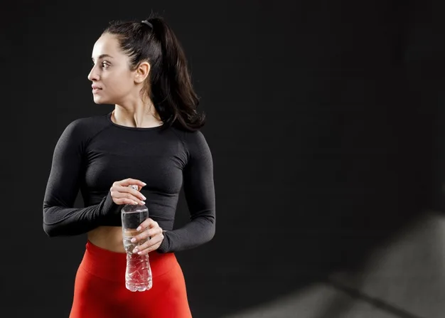 Mujer con ropa deportiva sosteniendo una botella de agua sobre fondo oscuro