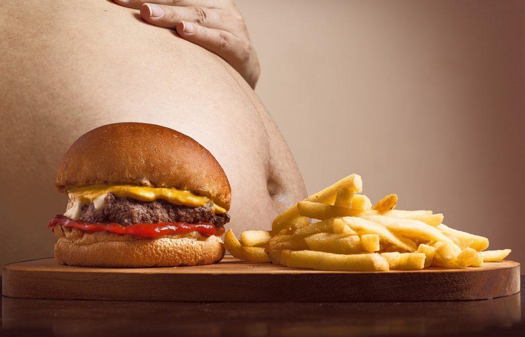 Hamburguesa y patatas fritas sobre una tabla, delante del abdomen prominente de una persona